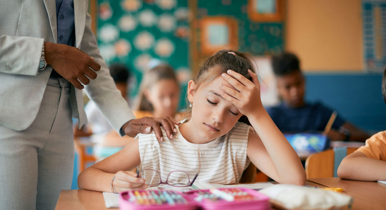 A young girl has a headache or fatigue while in the classroom, as her teacher comforts her.